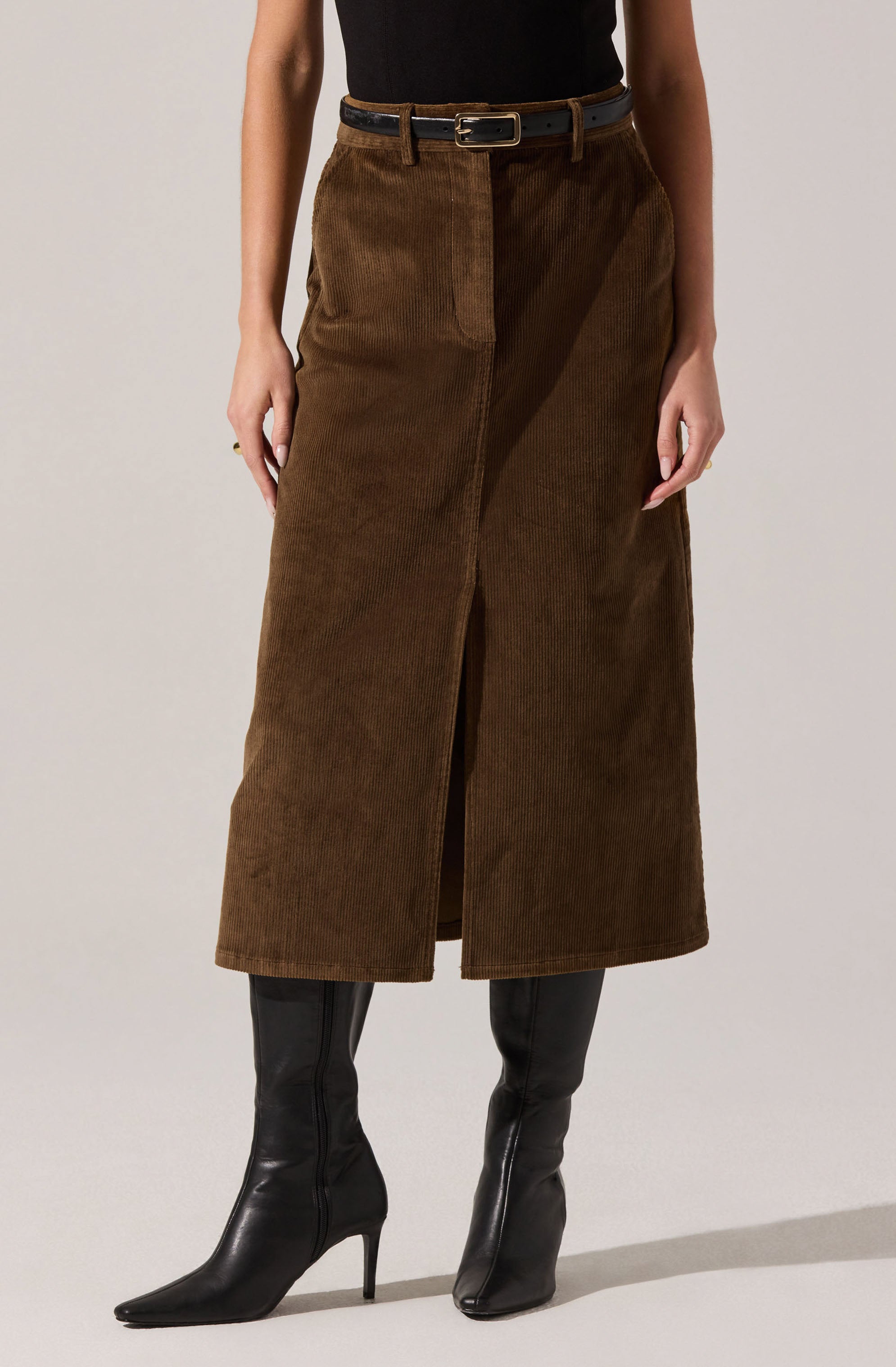 A woman is wearing a black sleeveless top tucked into a high-waisted, brown corduroy midi skirt with a front slit. She has a black belt with a gold buckle and is wearing black knee-high boots with pointed toes and high heels.