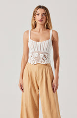 A woman is wearing a white crocheted tank top with wide straps and intricate openwork patterns, including floral and geometric designs. She pairs it with high-waisted, wide-leg beige trousers made from a lightweight fabric.