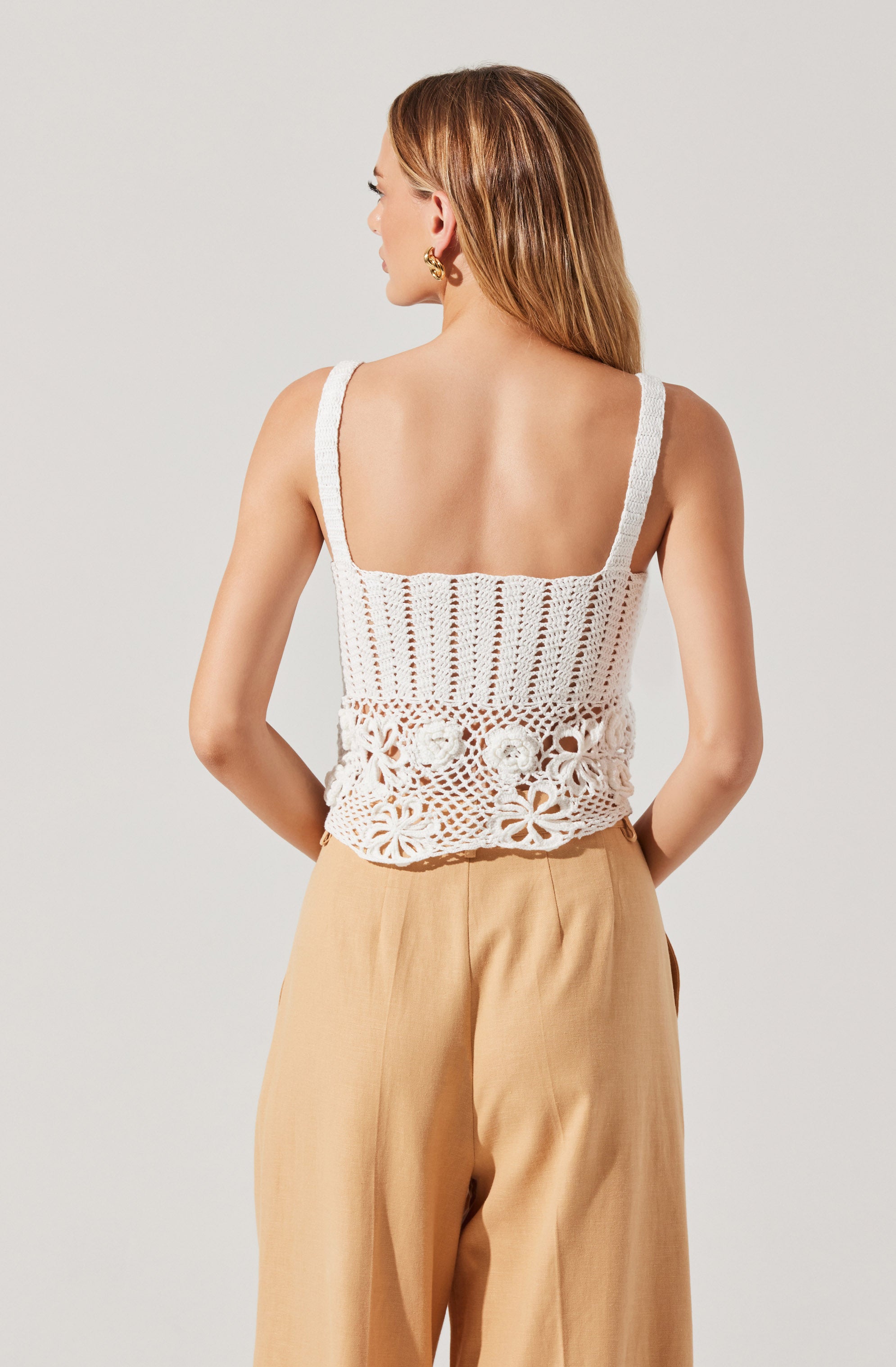 A woman is wearing a sleeveless, white crocheted top with wide straps and intricate open-knit patterns, including floral motifs at the hem. She is also wearing high-waisted, beige wide-leg pants.