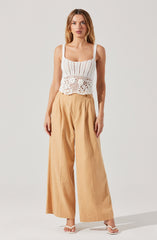A woman is wearing a sleeveless, white crocheted top with a fitted bodice and a scalloped hem. She pairs it with high-waisted, wide-leg beige pants that have pleats at the waist. She is also wearing white sandals with a small heel.