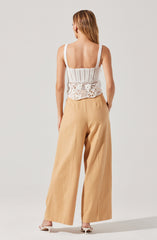 The woman is wearing a white sleeveless knit tank top with straps, featuring a crochet-style pattern with floral and geometric designs. She pairs it with high-waisted, wide-leg beige pants.