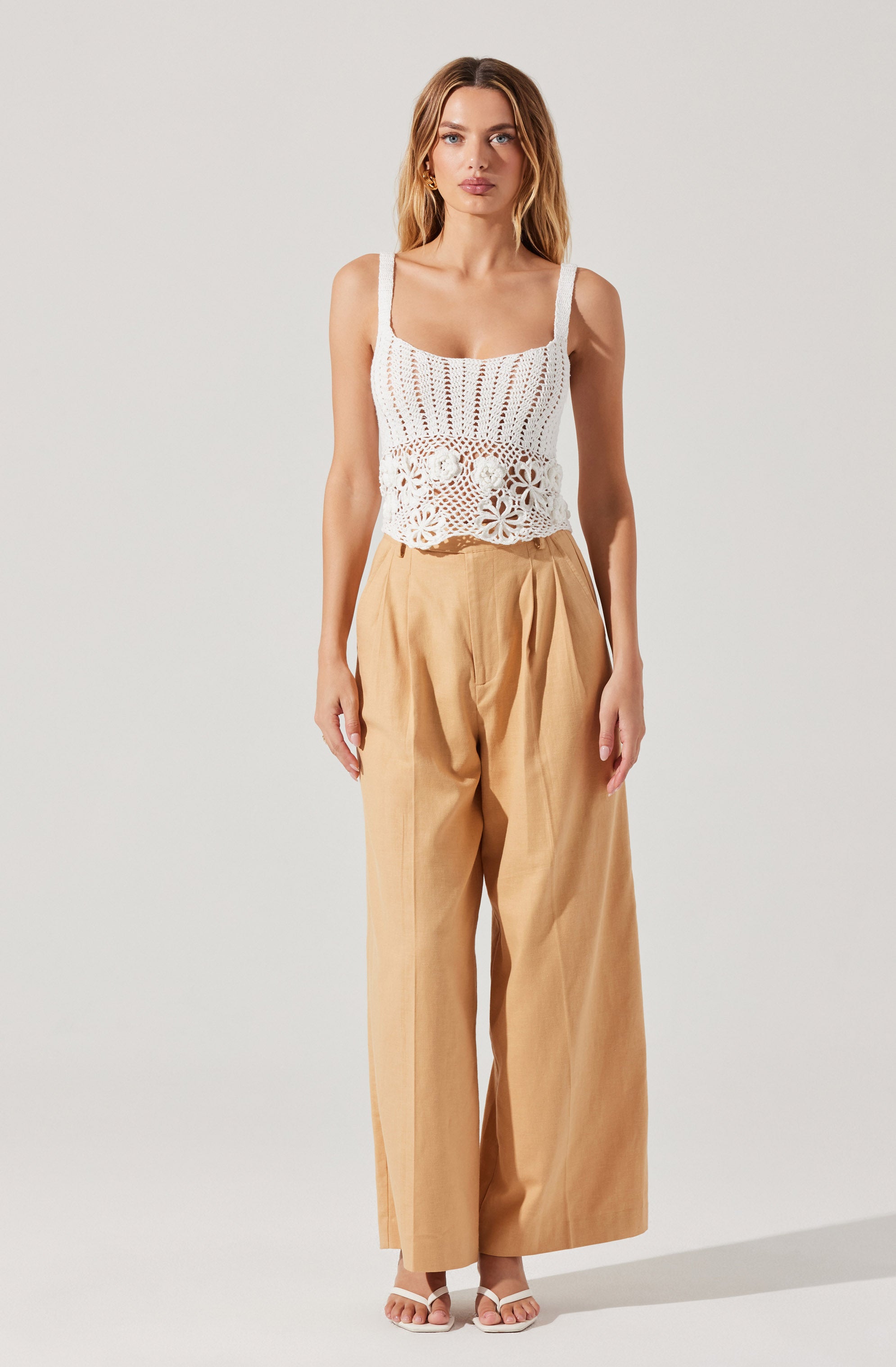 A woman is wearing a white crochet tank top with wide straps, featuring a floral pattern at the bottom edge. She pairs it with high-waisted, wide-leg beige trousers. She is also wearing white open-toe sandals.