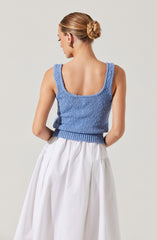 A woman is wearing a sleeveless, light blue knitted top with wide shoulder straps and a scoop neckline. She pairs it with a high-waisted, white, flowing skirt that reaches below her knees. Her hair is styled in a neat twisted bun at the back of her head.