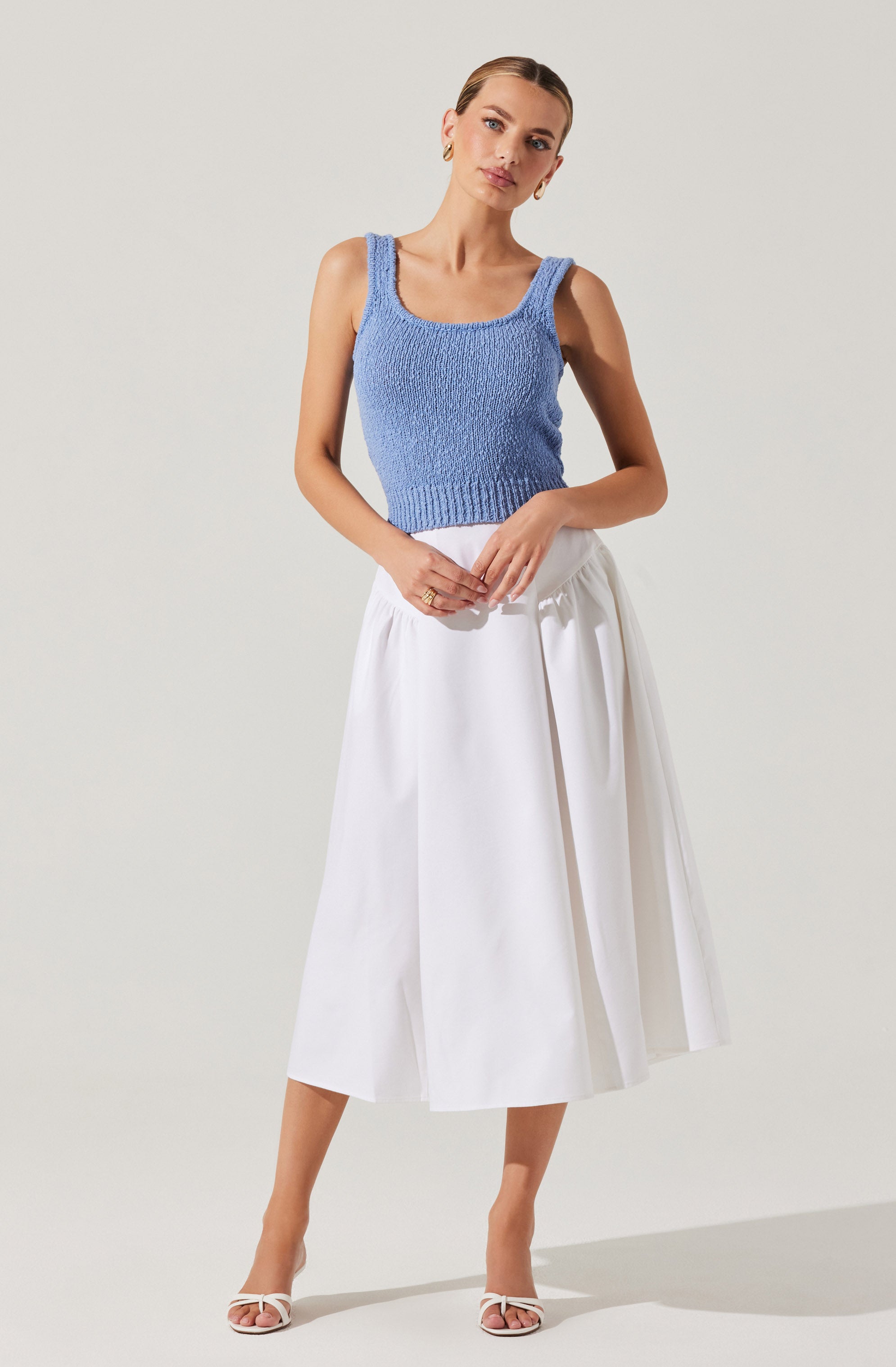 A woman is wearing a sleeveless, light blue knit top with wide straps and a rounded neckline. She pairs it with a high-waisted, white midi skirt that features pleats and a tie detail at the waist. She completes her outfit with strappy white heels.