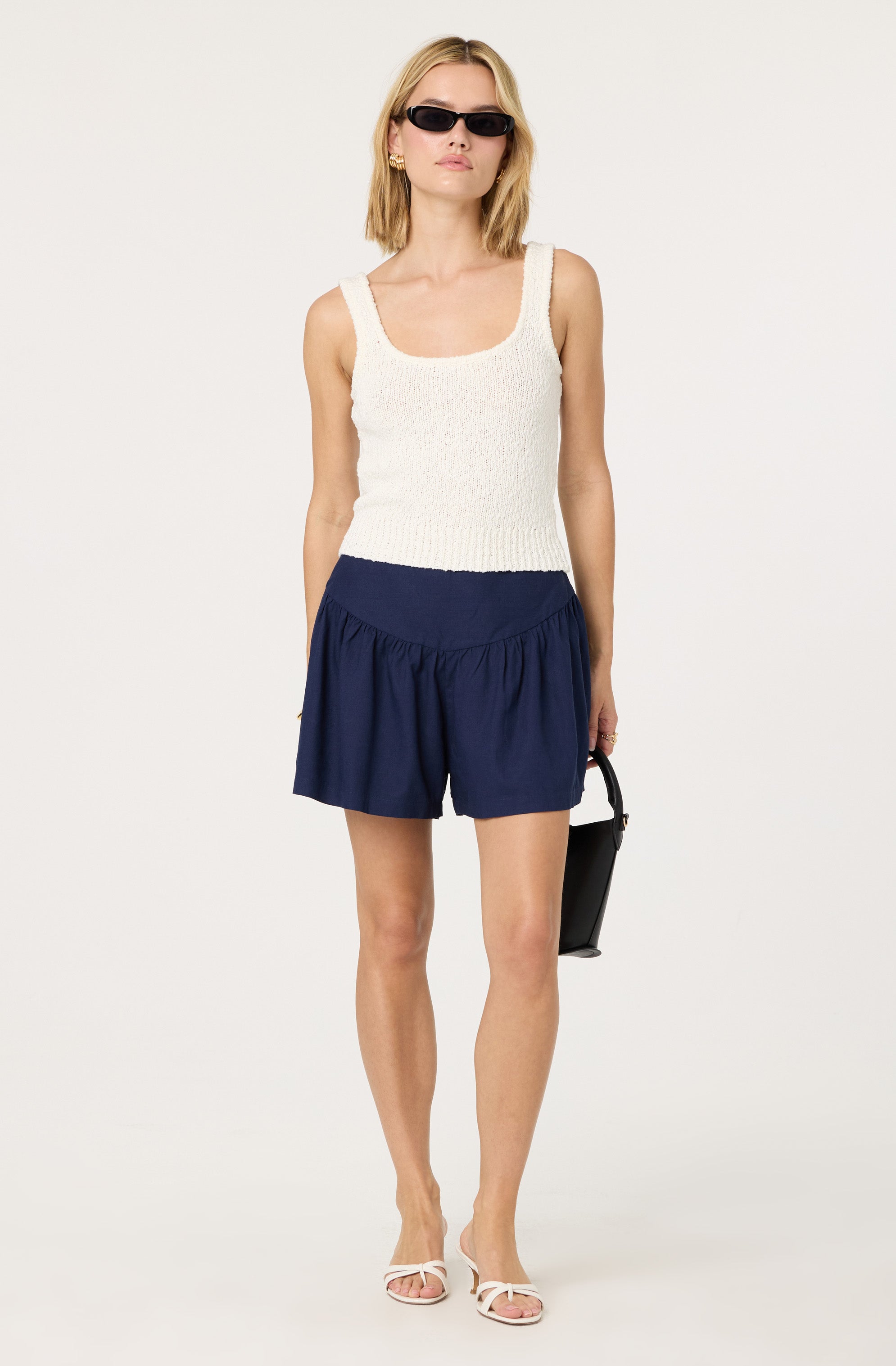 A woman is wearing a sleeveless, cream-colored, textured knit tank top with wide shoulder straps and a scoop neckline. She pairs it with high-waisted, navy blue shorts that have a loose fit and gathered fabric at the waist. She is also wearing white strappy sandals, black rectangular sunglasses, and gold earrings, and she is holding a small black handbag.