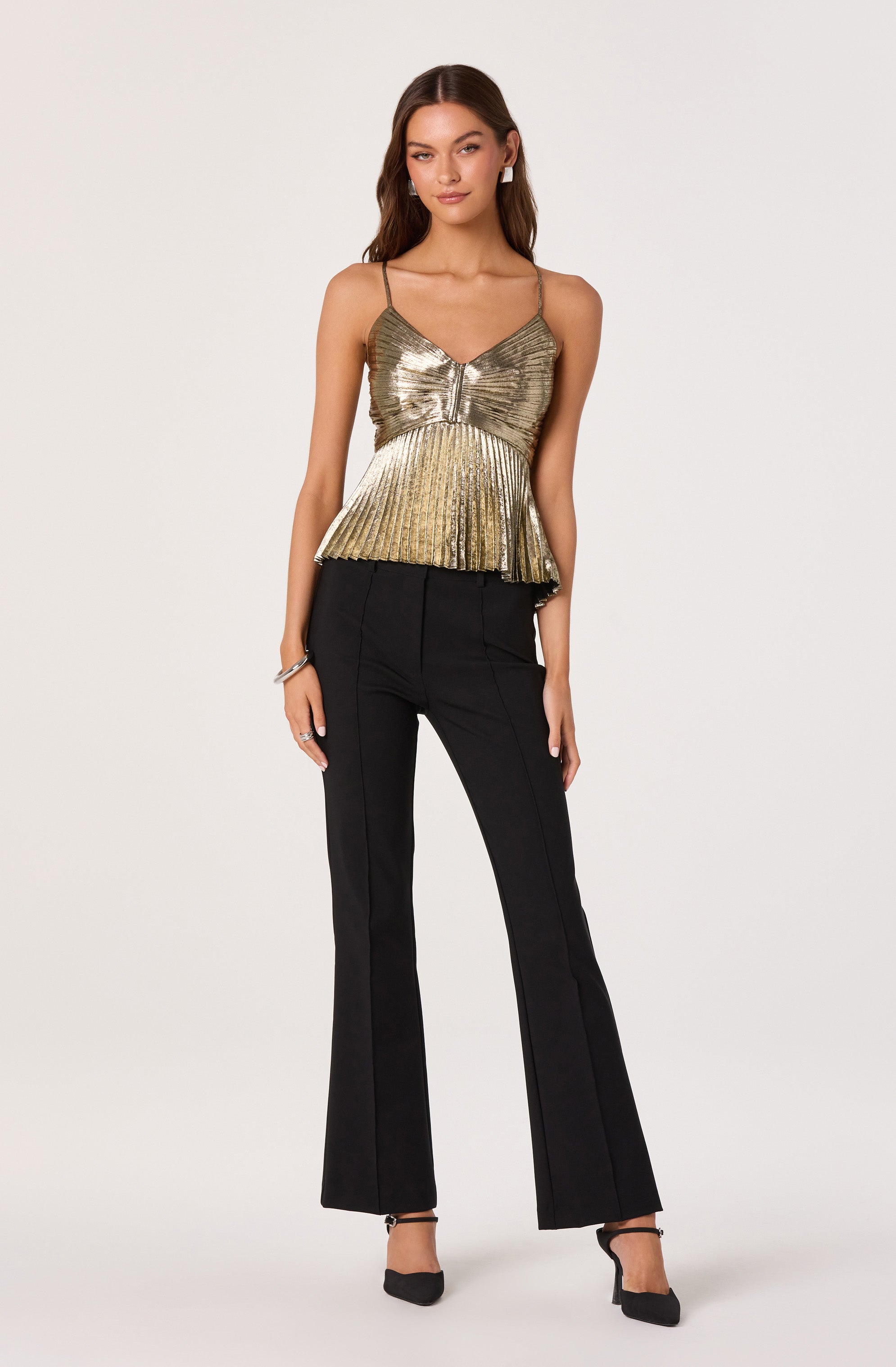 A woman is wearing a gold metallic sleeveless top with thin spaghetti straps, a ruched detail at the center, and a ruffled hemline. She pairs the top with high-waisted, black, wide-leg trousers that have front seams and a tailored fit. She completes the look with black ankle-strap heels.