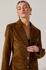 A woman is wearing a brown corduroy blazer with a notched lapel collar, two front flap pockets, and two buttons down the front. She has earrings with large, gold-colored disc and purple bead accents, and a gold ring on her right hand.