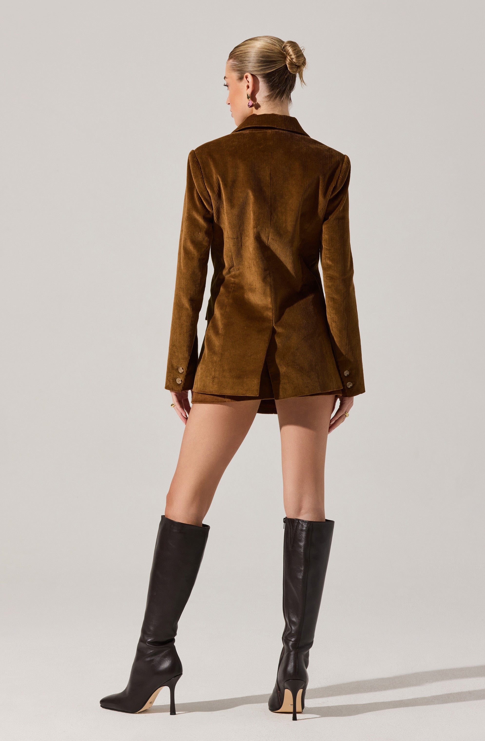 A woman is wearing a brown velvet blazer with long sleeves that have three buttons on each cuff. She is also wearing tall black leather high-heeled boots that reach just below her knees.