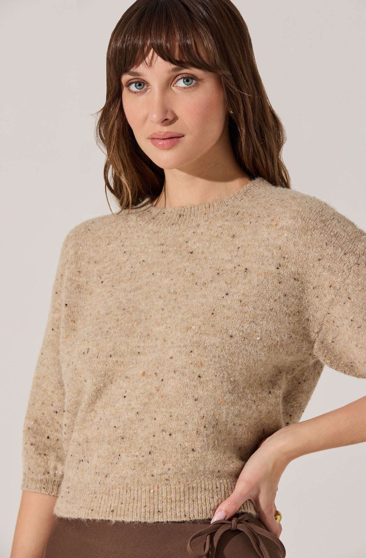 A woman is wearing a beige, speckled knit sweater with a crew neckline and three-quarter length sleeves. The sweater has a soft, textured appearance with small multicolored specks throughout.