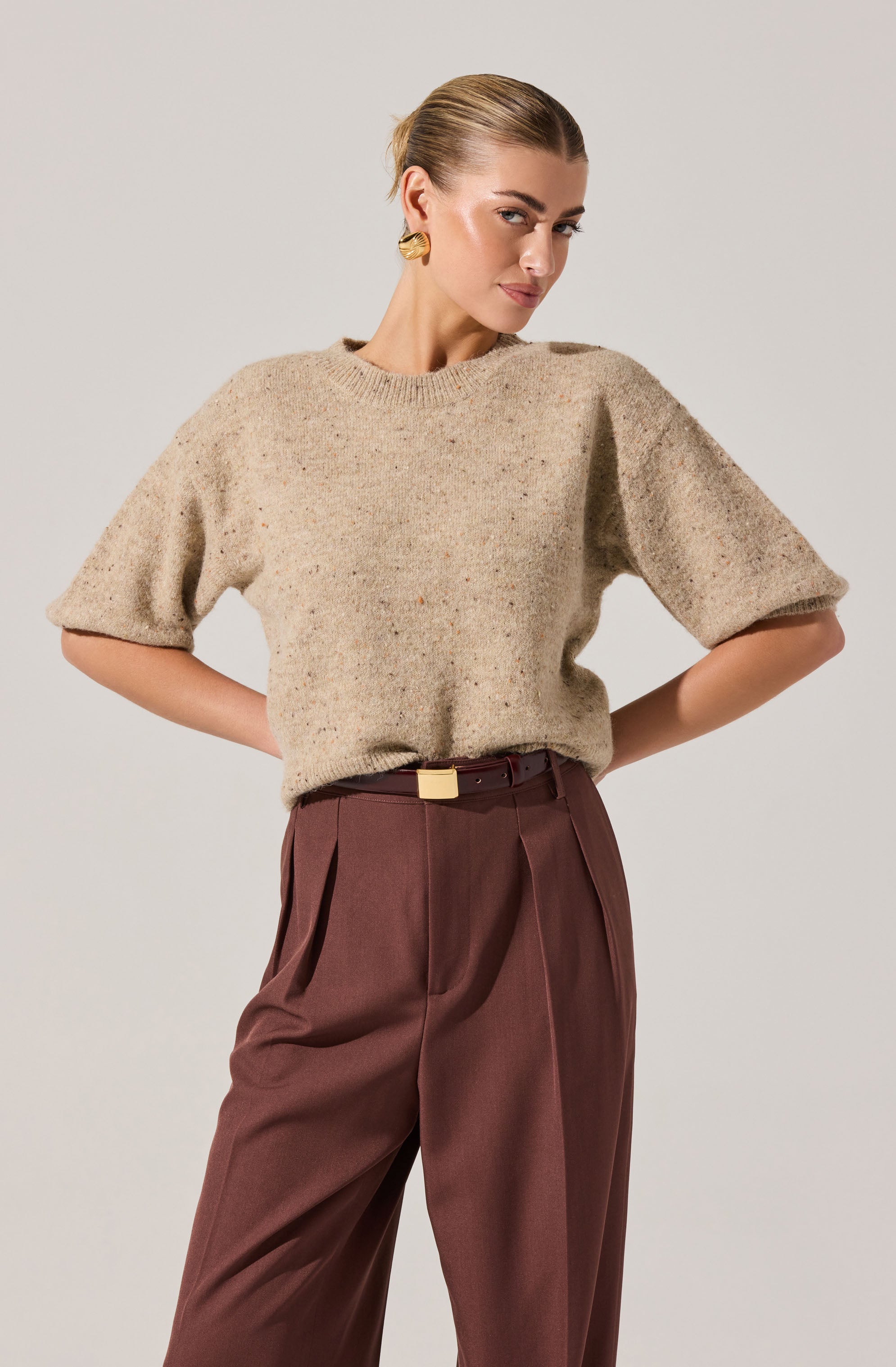 A woman is wearing a beige, short-sleeved, slightly oversized knit sweater with a rounded neckline, paired with high-waisted, pleated, brown trousers that have a belt with a rectangular buckle. She has her hands on her hips, and her hair is styled in a sleek, pulled-back look. She is accessorized with large, gold, sculptural earrings.