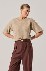 A woman is wearing a beige, short-sleeved, slightly oversized knit sweater with a rounded neckline, paired with high-waisted, pleated, brown trousers that have a belt with a rectangular buckle. She has her hands on her hips, and her hair is styled in a sleek, pulled-back look. She is accessorized with large, gold, sculptural earrings.