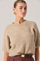 A woman is wearing a beige, fuzzy, short-sleeved sweater with a round neckline. She is also dressed in high-waisted, dark red pants with a wide belt featuring a square gold buckle. She has gold, textured, round earrings.