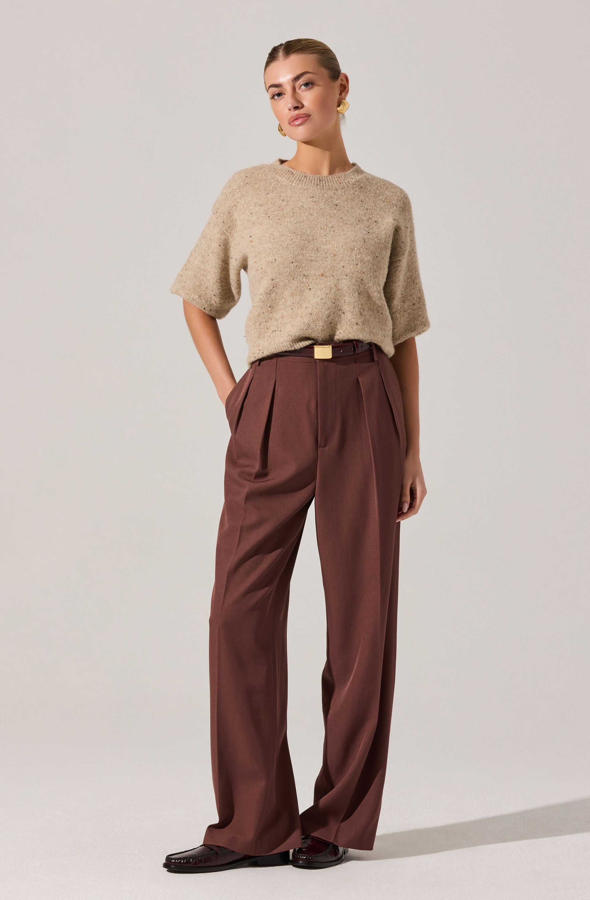 A woman is wearing a loose, beige, textured short-sleeve sweater with a round neckline, tucked into high-waisted, wide-leg brown trousers with pleats at the waist. She has a small belt with a rectangular gold buckle, and she is wearing dark brown shoes featuring a slight heel. She also has yellow earrings and is standing against a plain light background.
