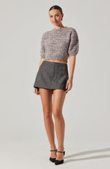 A woman is wearing a loose-fitting, textured, cropped sweater with three-quarter puff sleeves in shades of beige and light brown. She pairs it with a high-waisted, gray plaid mini skirt that has a tailored fit. She is also wearing black ankle-strap high heel shoes.