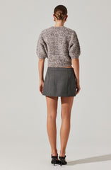 A woman is wearing a textured, short-sleeved sweater with a rounded neckline in shades of gray and white. She pairs it with a high-waisted, gray plaid mini skirt that has subtle checkered patterns. She completes the outfit with black strappy high-heeled shoes.
