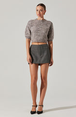 The woman is wearing a cropped, loose-fitting sweater with a fuzzy, textured material in shades of gray and beige. She is also wearing a high-waisted, dark gray, plaid mini skirt with subtle checkered pattern and front pockets. She has on black high-heeled shoes with ankle straps and gold hoop earrings.