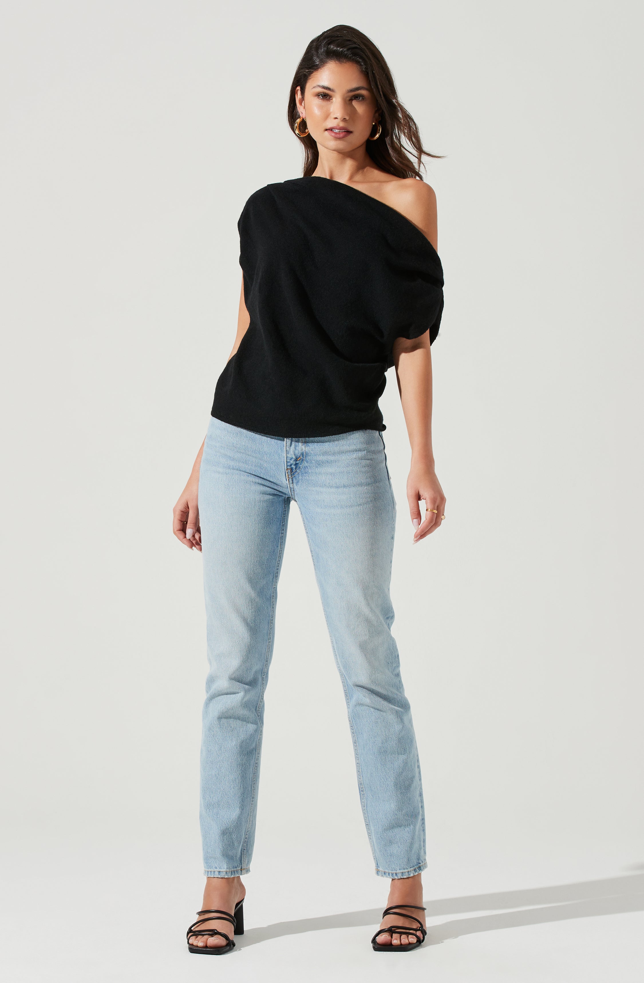 A woman is wearing a black, oversized, off-the-shoulder sweater with voluminous, puffed sleeves, paired with light-wash, high-waisted straight-leg jeans and black strappy high-heeled sandals. She has gold hoop earrings and a subtle makeup look.