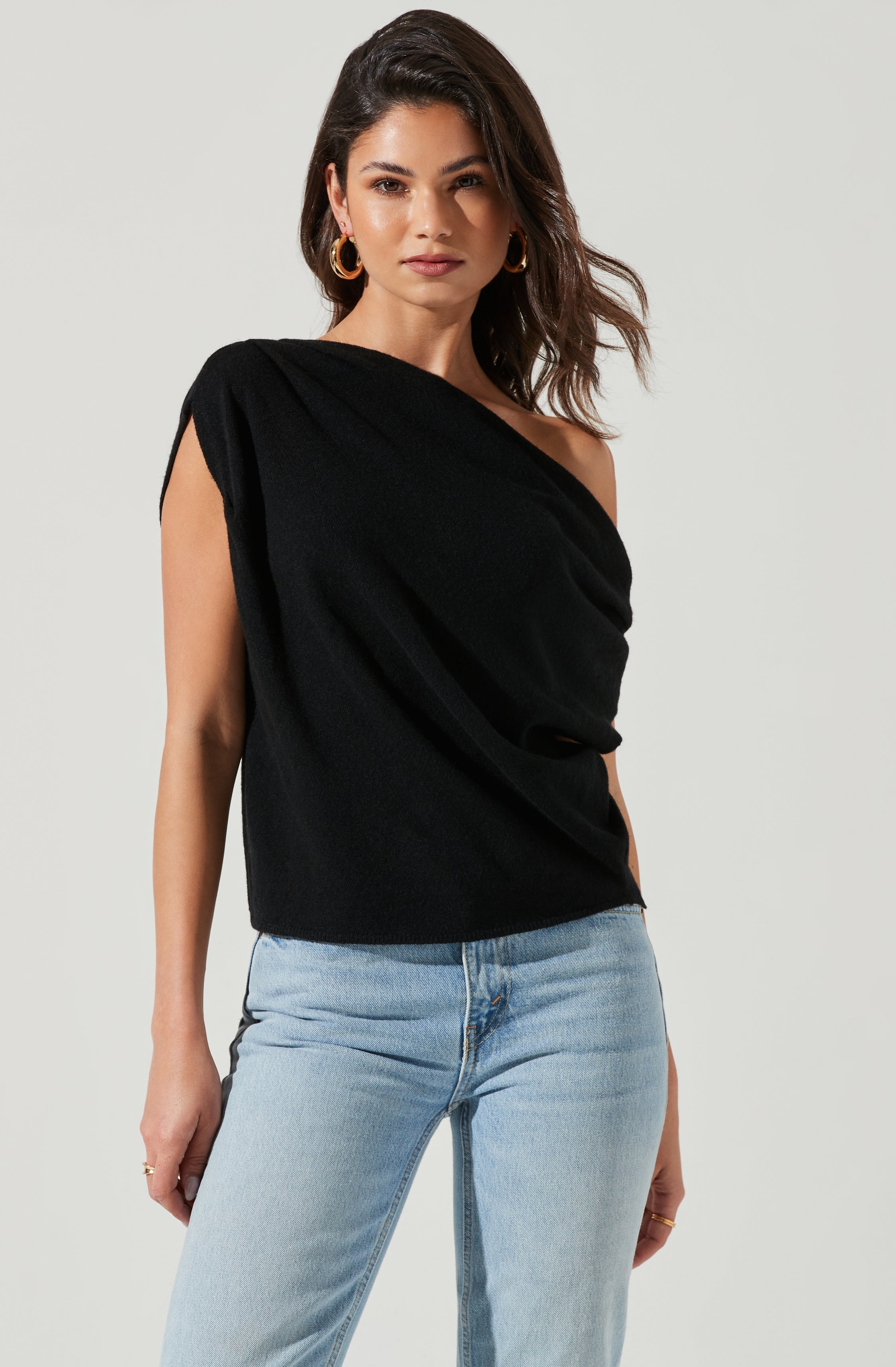 A woman is wearing a black sleeveless top with a draped, asymmetrical design that exposes one shoulder. She is also wearing light-washed jeans.