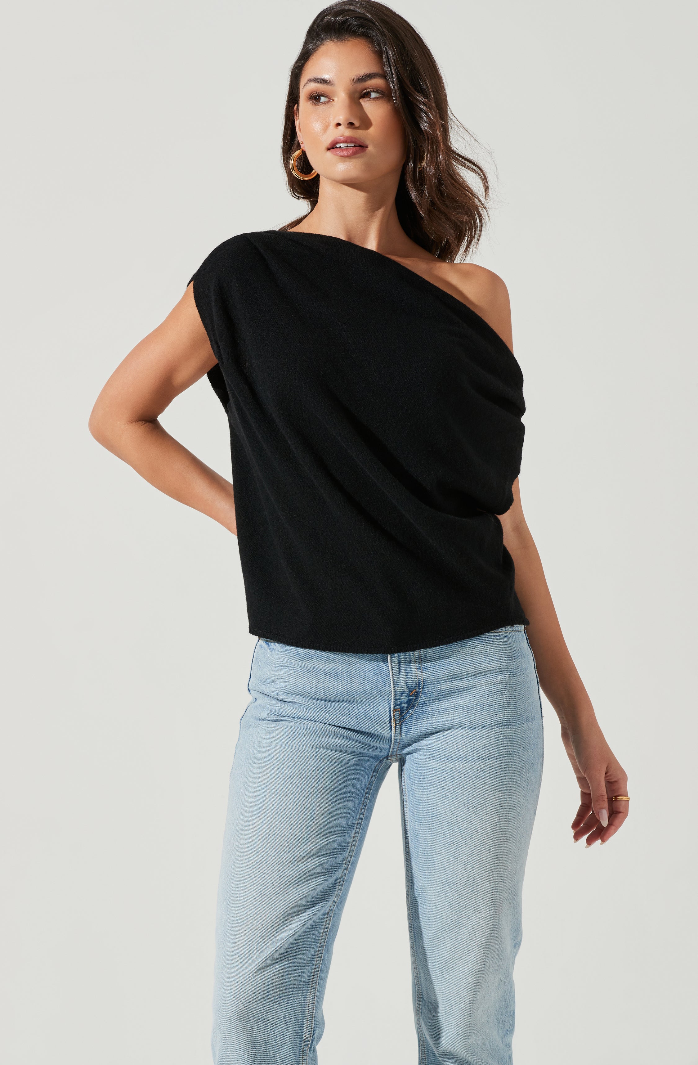 A woman is wearing a black, asymmetrical, one-shoulder top with a loose fit, paired with light blue denim jeans. The top drapes across one shoulder, exposing the other shoulder, and has a relaxed, slightly textured fabric.