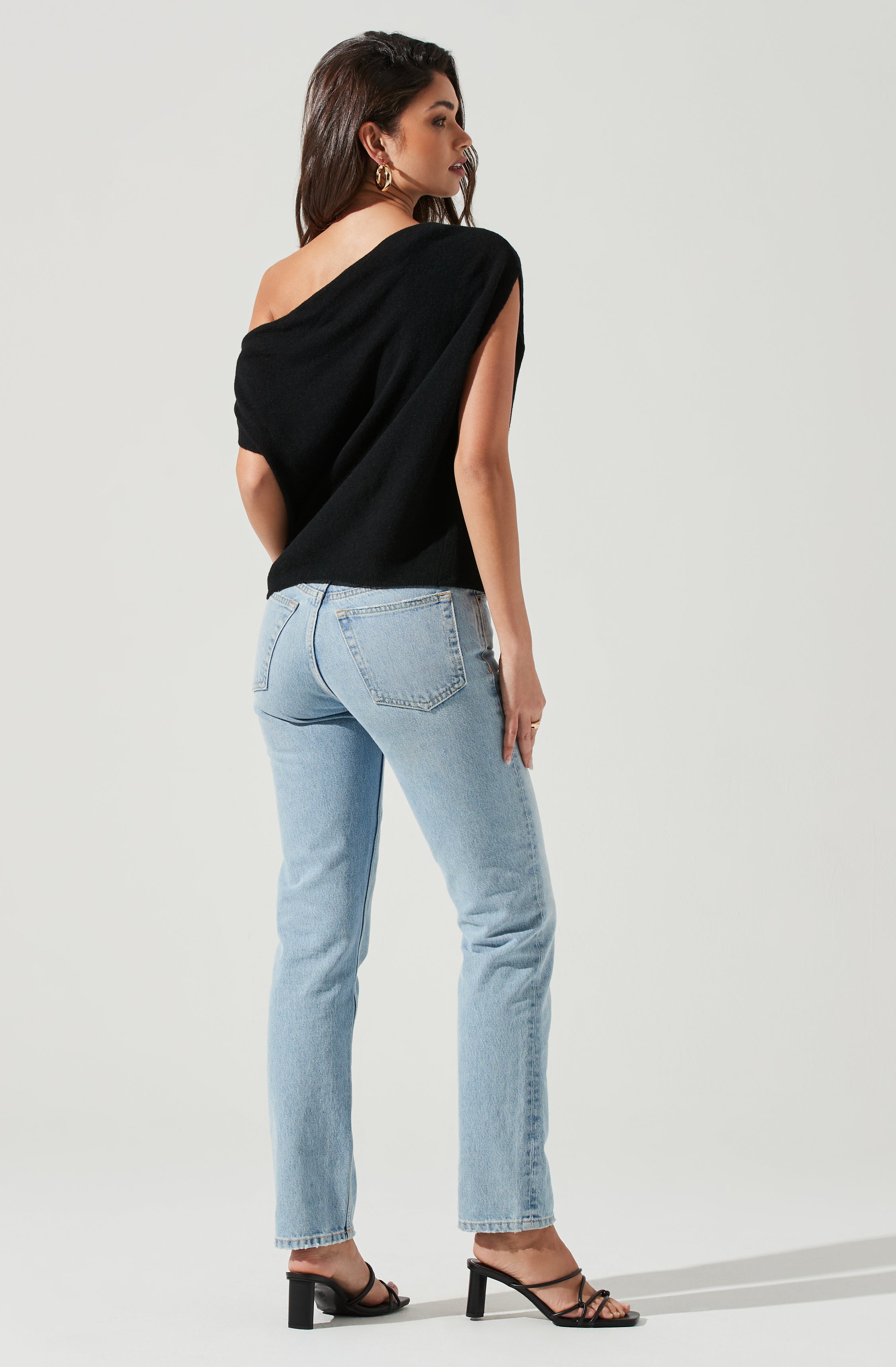 A woman is wearing a black sleeveless top with an off-the-shoulder design and light blue high-waisted jeans. She has accessorized with large gold hoop earrings and is wearing black high-heeled sandals with thin straps.