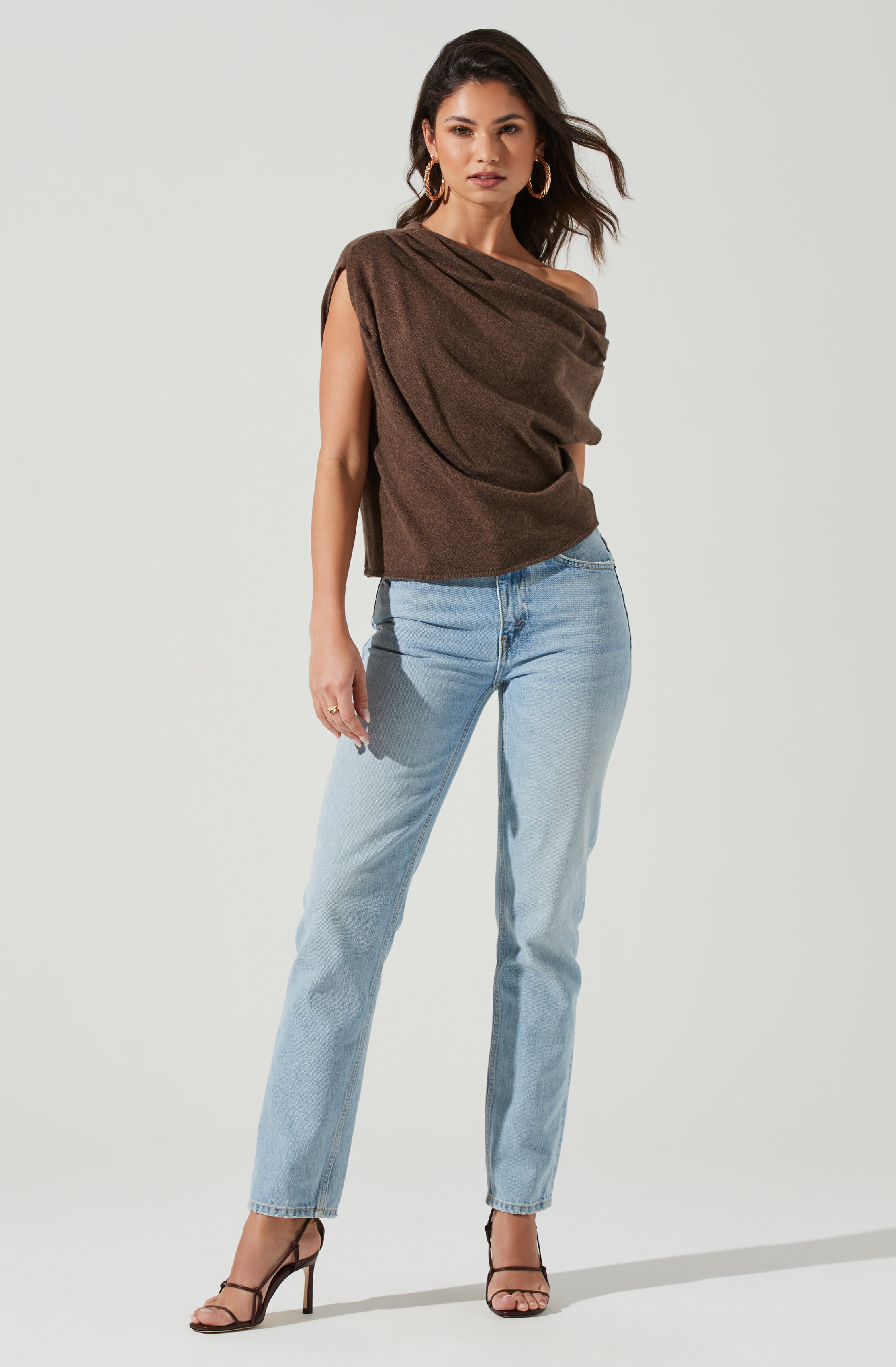A woman is wearing a loose, off-the-shoulder, dark brown top made from a soft, textured fabric. She pairs it with light blue, high-waisted jeans that have a relaxed fit. Her outfit is completed with black, open-toe high-heeled sandals featuring thin ankle straps. She has accessorized with large hoop earrings.