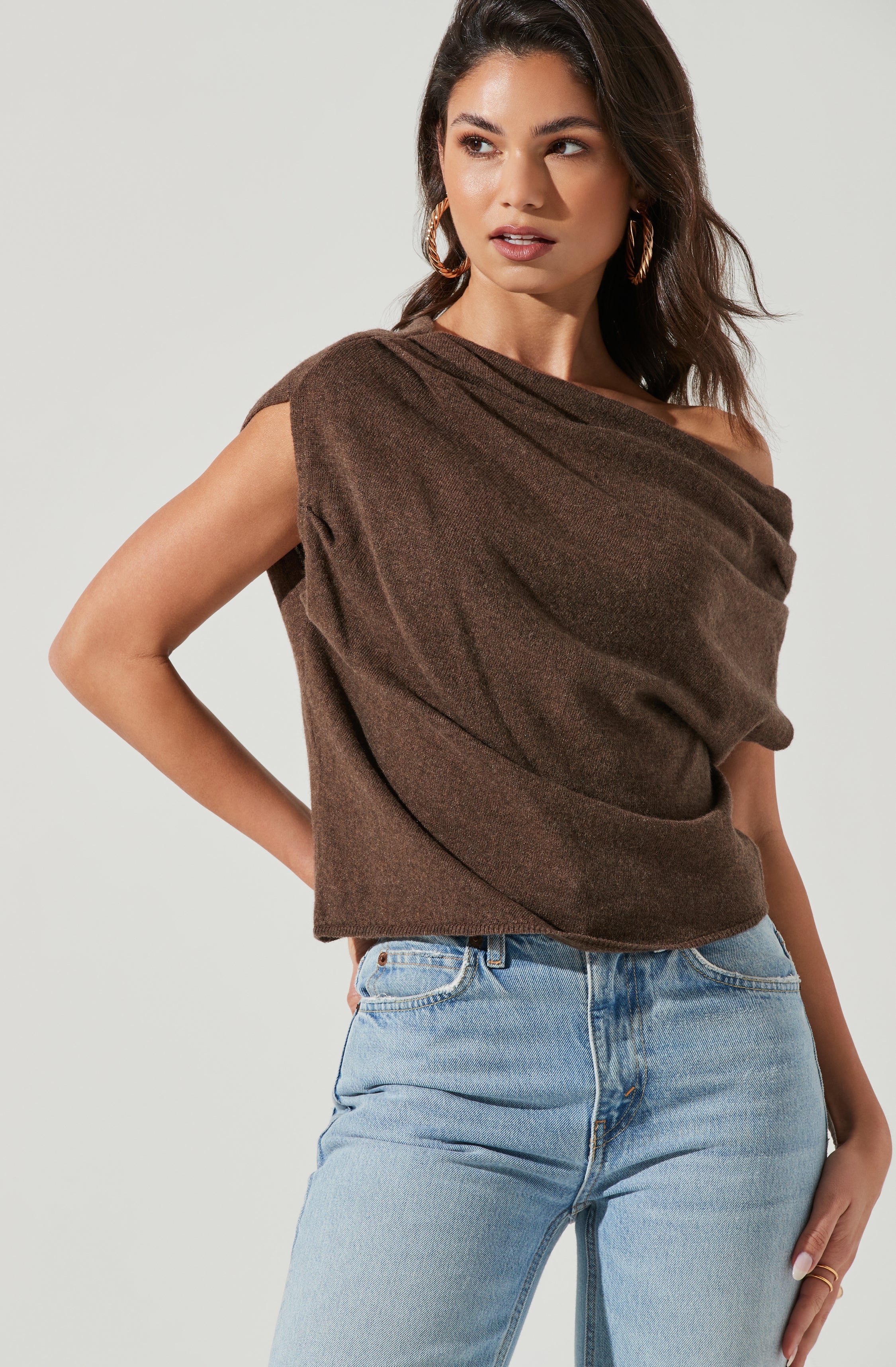 A woman is wearing a loose-fitting, brown sleeveless top with a draped neckline and light blue high-waisted jeans.
