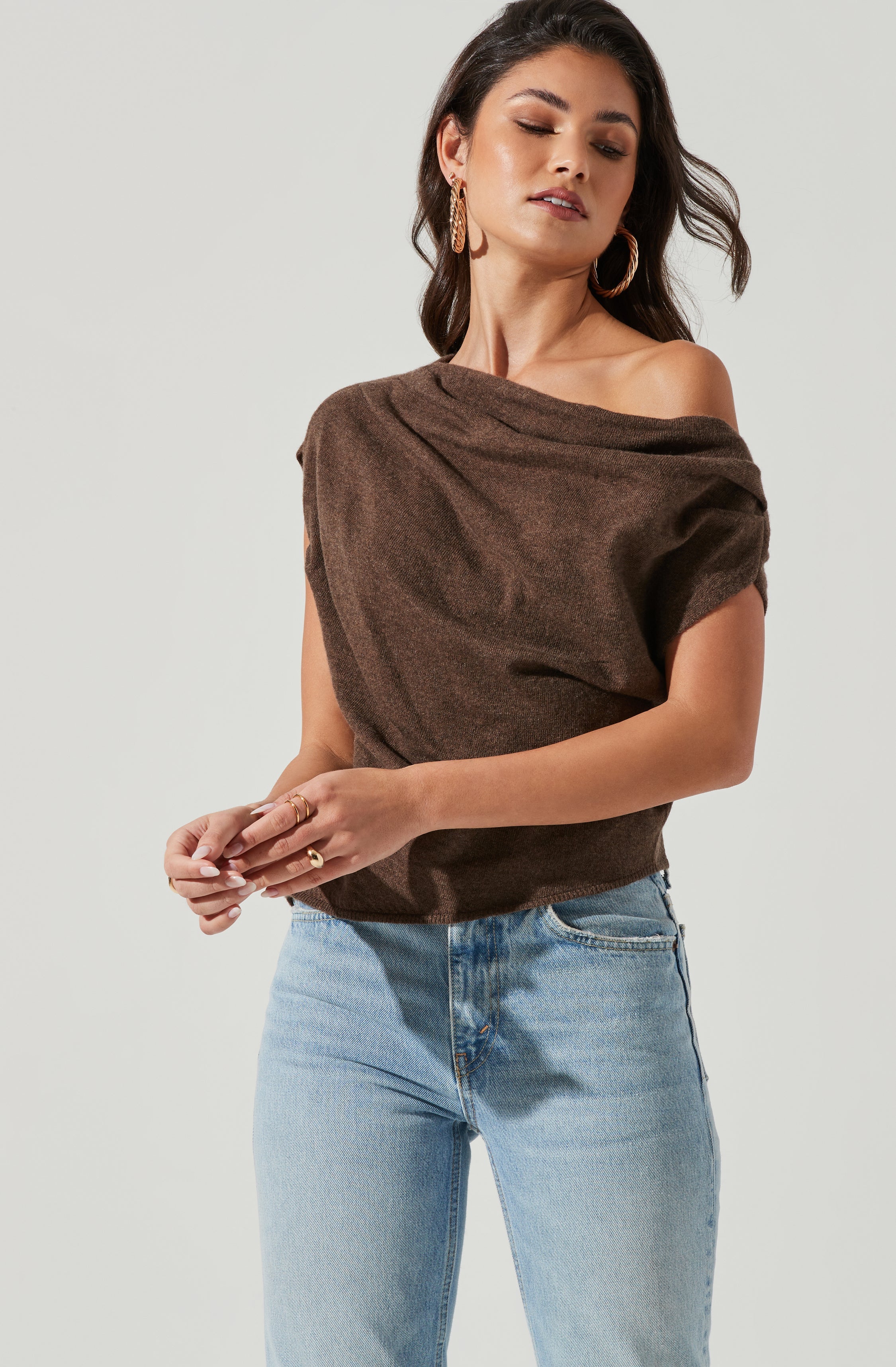 A woman wearing a dark brown, off-the-shoulder top with a relaxed fit and rolled sleeves, paired with light blue high-waisted jeans. She accessorizes with large gold hoop earrings and multiple rings on her fingers.