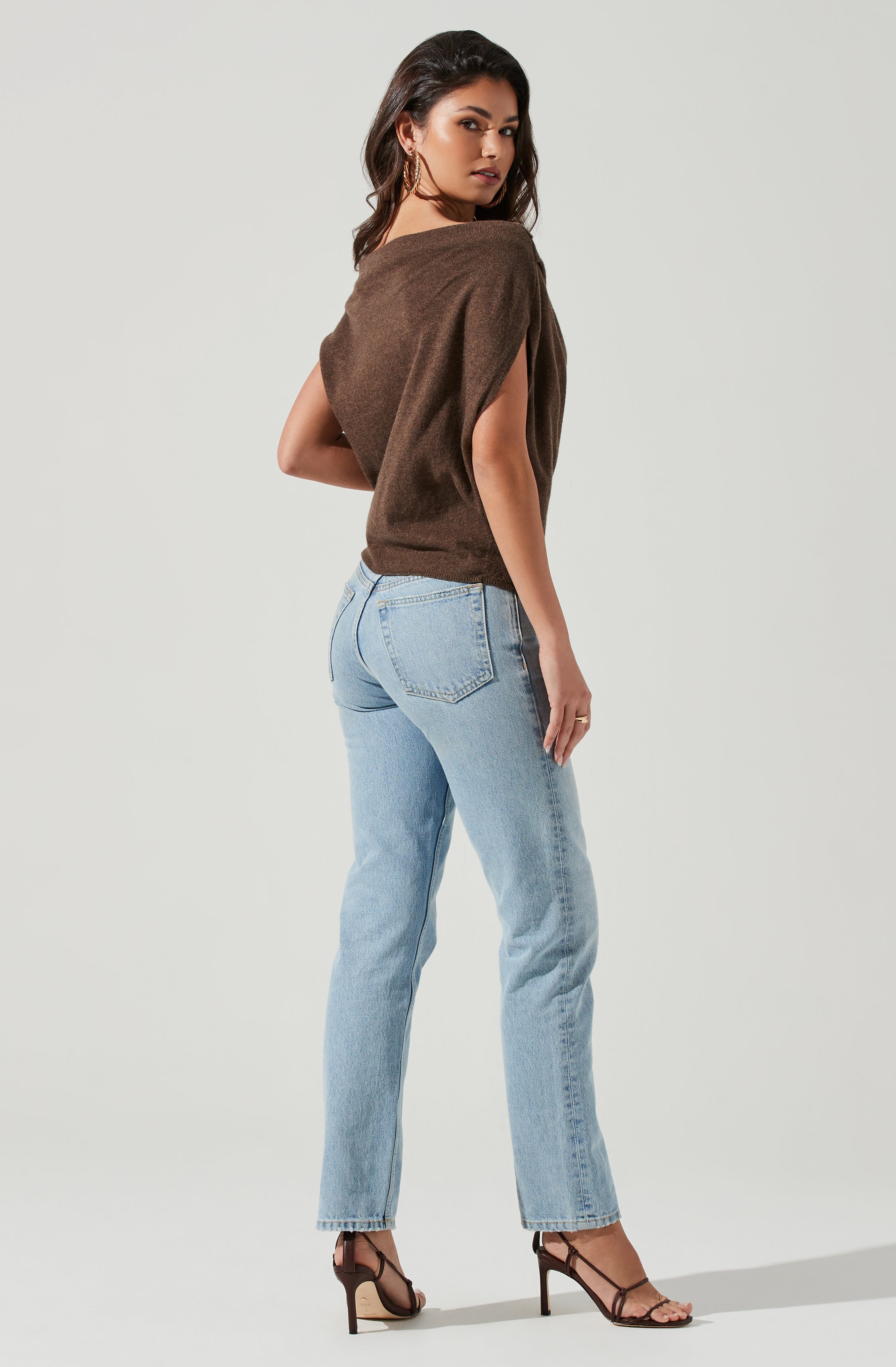 A woman is wearing a sleeveless, brown sweater with a loose fit and a wide neckline. She is also wearing light blue, high-waisted jeans with a classic five-pocket design. Her footwear consists of black, strappy high-heeled sandals.