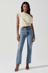 A woman is wearing a sleeveless, light yellow, draped top that exposes one shoulder. She has paired it with high-waisted, light blue jeans and a black belt with a gold circular buckle. She is also wearing black high-heeled sandals and accessorized with gold hoop earrings and rings.