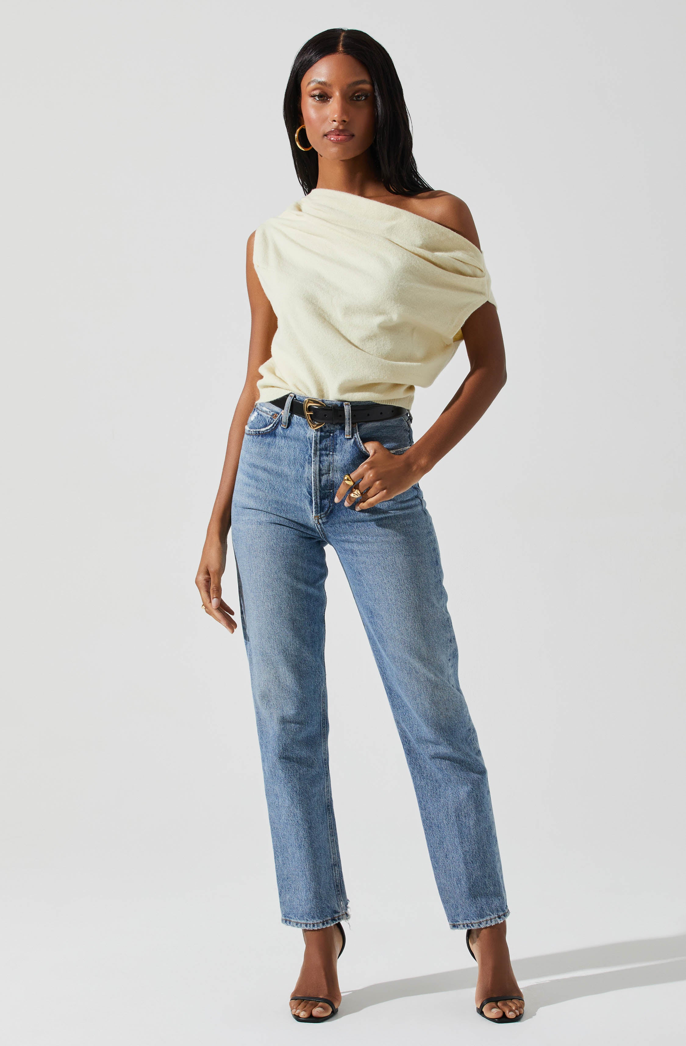 A woman is wearing a cream-colored, off-the-shoulder top with a draped, loose fit. She pairs it with high-waisted, light-wash jeans and a black belt with a gold buckle. She accessorizes with gold hoop earrings and large rings. She is also wearing black high-heeled sandals.