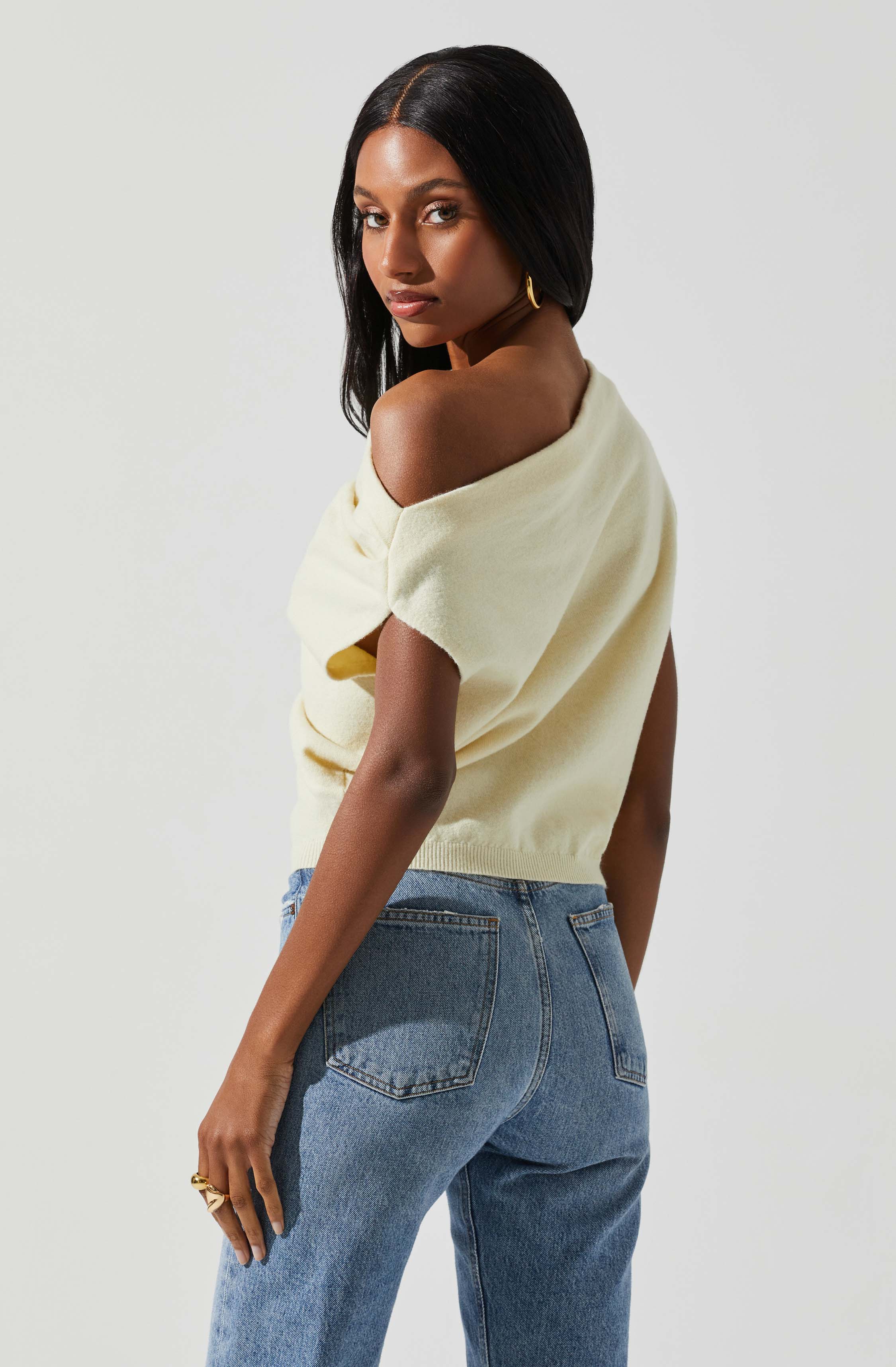 A woman is wearing a light yellow, off-the-shoulder sweater and high-waisted blue jeans.