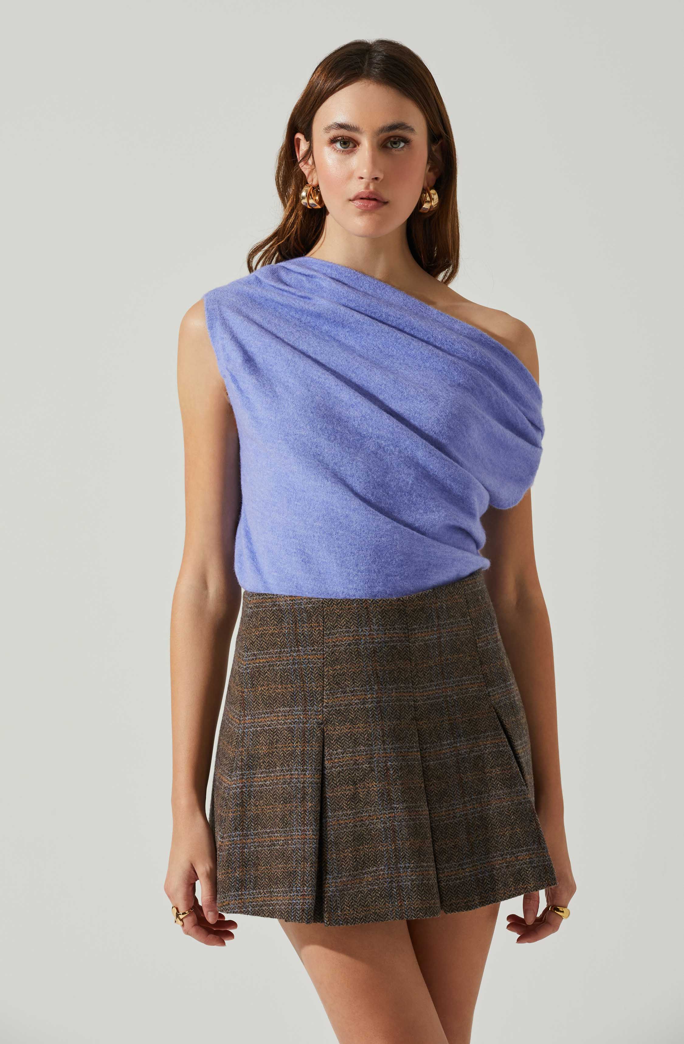 A woman is wearing a sleeveless, draped, light purple top with a one-shoulder design, paired with high-waisted, brown plaid shorts featuring pleats. She accessorizes with gold hoop earrings and rings on her fingers.