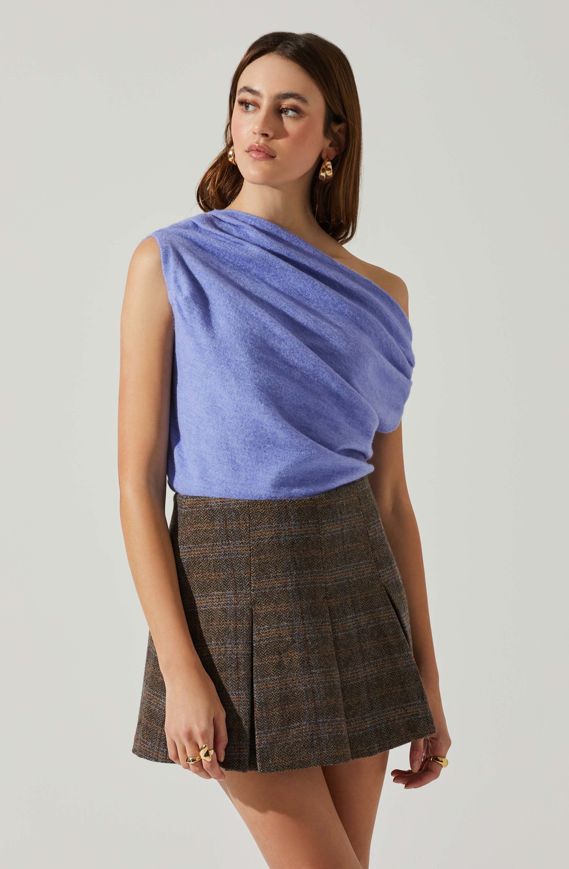A woman is wearing a sleeveless, asymmetrical, lavender-colored top made of a textured, soft fabric. She pairs it with high-waisted, brown plaid pleated shorts. She accessorizes with gold hoop earrings and multiple rings.