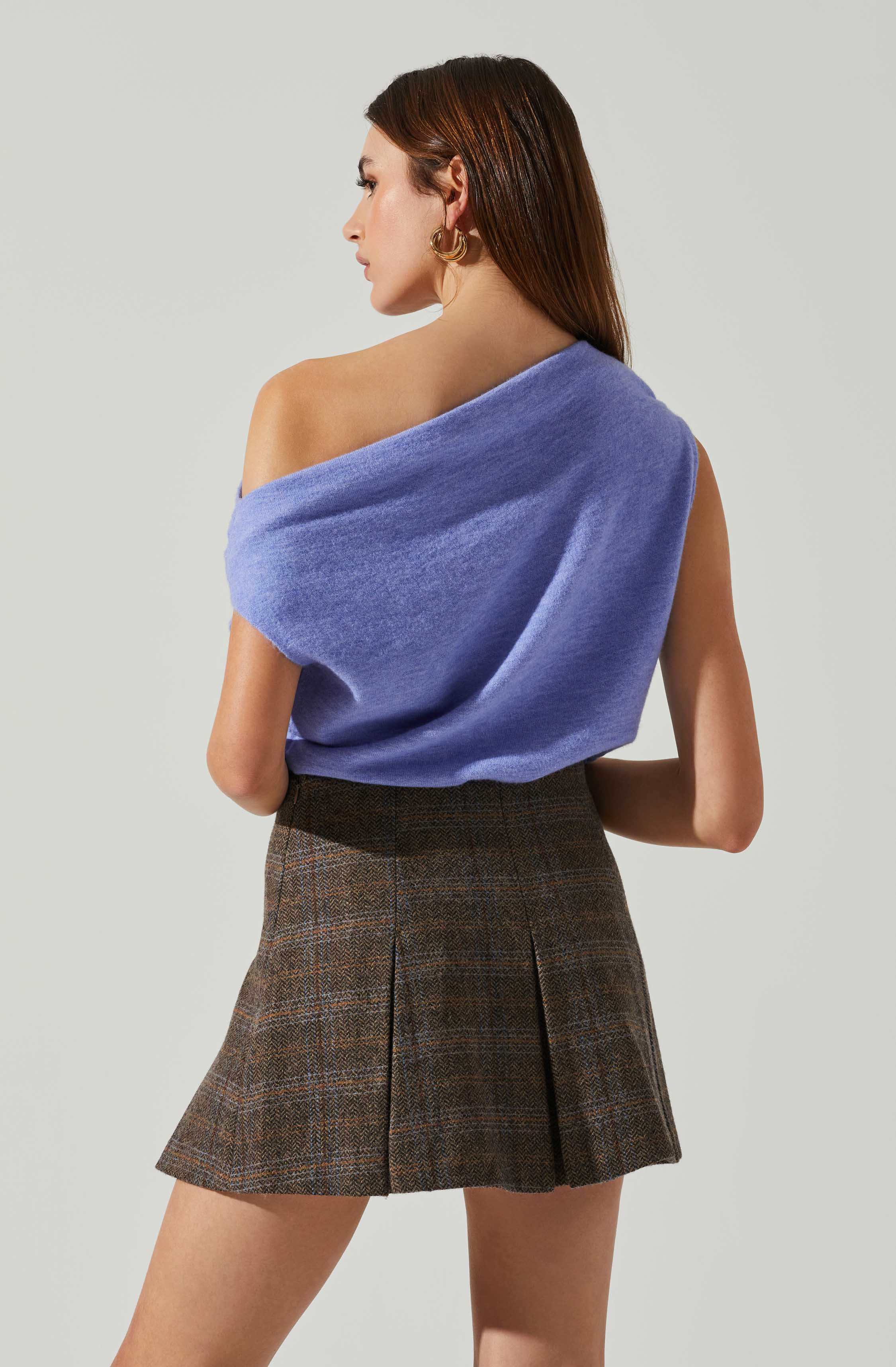 A woman is wearing a soft, powder-blue, off-the-shoulder top made of a plush, fuzzy fabric. She pairs it with a high-waisted, brown plaid skirt featuring pleats and a subtle checkered pattern. She has long, straight, brown hair and is accessorized with large gold hoop earrings.