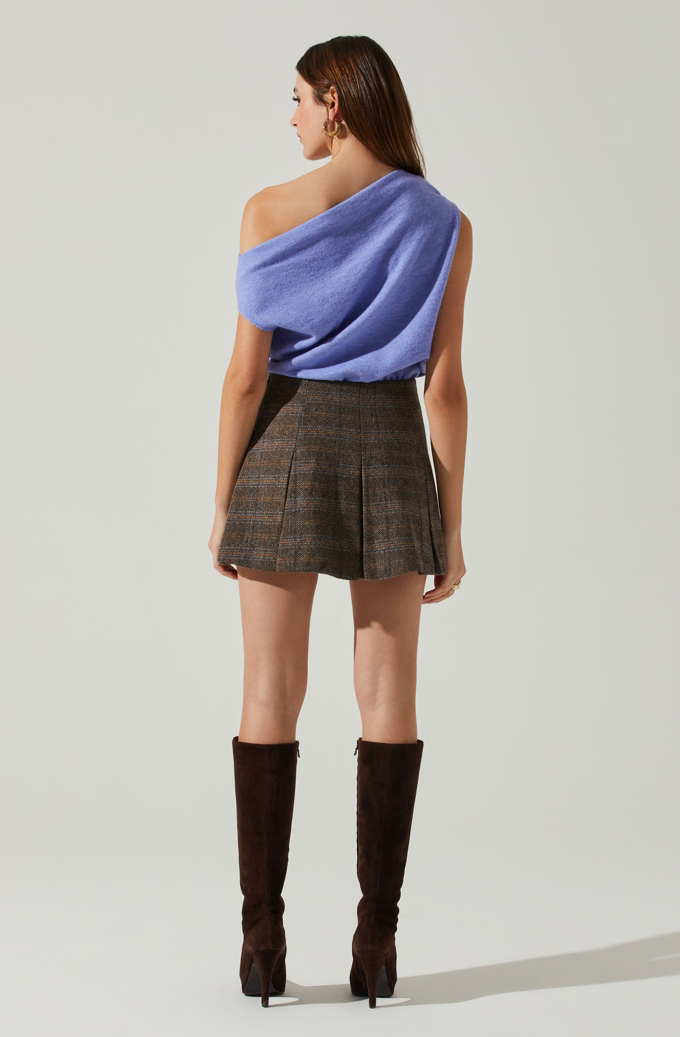 A woman is wearing an off-the-shoulder, sleeveless, light purple top made of a soft, fuzzy material. She pairs it with a high-waisted, brown plaid mini skirt that features pleats. She is also wearing knee-high, dark brown suede boots with high heels.