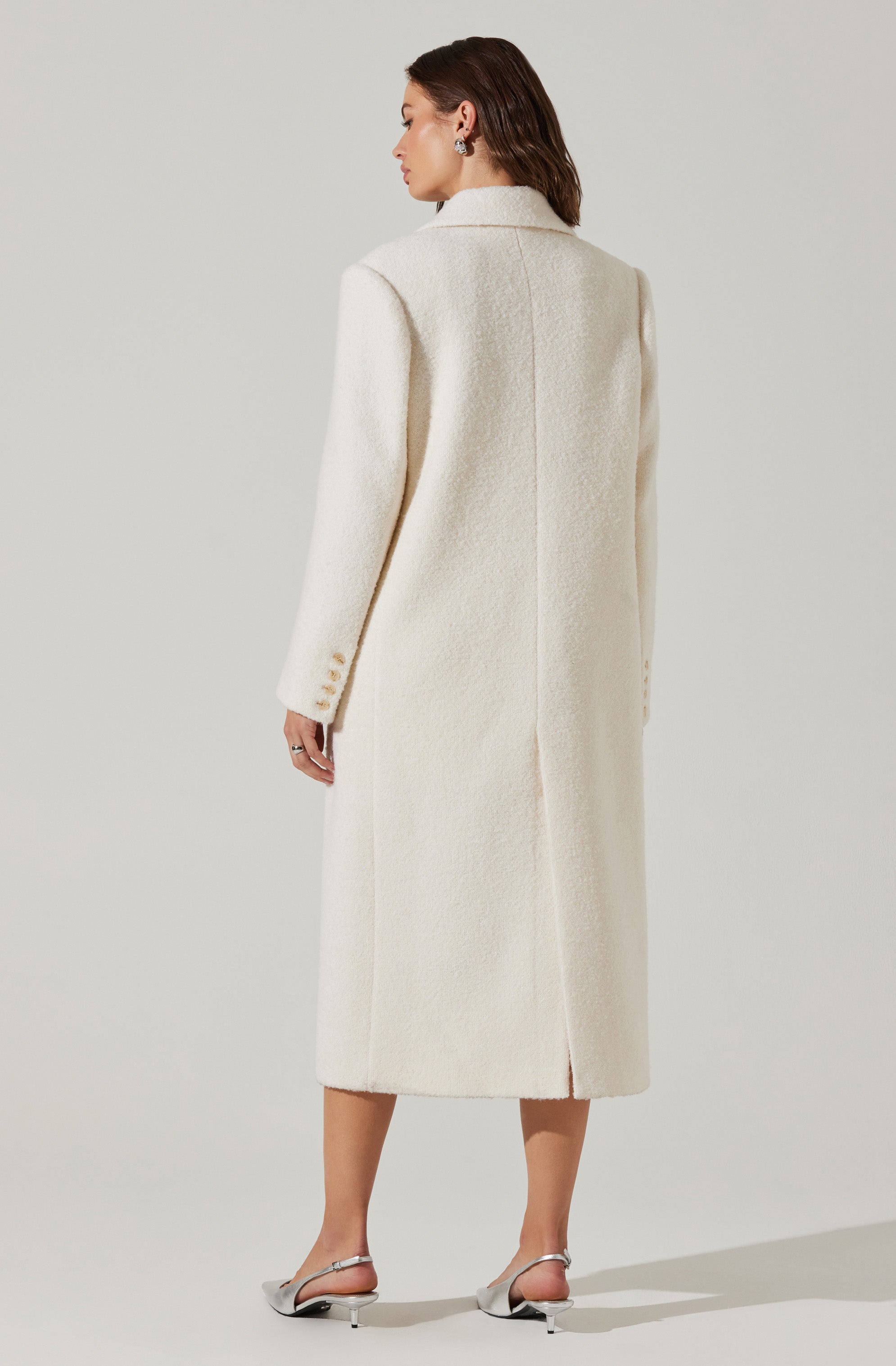 The woman is wearing a long, cream-colored coat made of a textured, possibly boucle or wool material. The coat features a notched lapel collar, and the back has a center seam with a slit at the hem. Her left sleeve has three small buttons near the cuff. She is also wearing silver, open-toed heels with ankle straps.