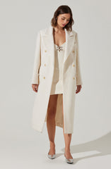A woman is wearing a long, white double-breasted coat with large buttons, paired with a white dress that has a lace-up neckline. She is also wearing silver high-heeled shoes.