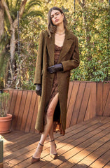 A woman is wearing an elegant, long brown coat with a double-breasted design and wide lapels. Underneath, she has on a brown dress with a slit that reveals her leg. She is also wearing black gloves and brown high-heeled shoes with ankle straps. Accessories include a gold necklace and hoop earrings. She has shoulder-length brown hair and is standing on a wooden deck outdoors, surrounded by green foliage.