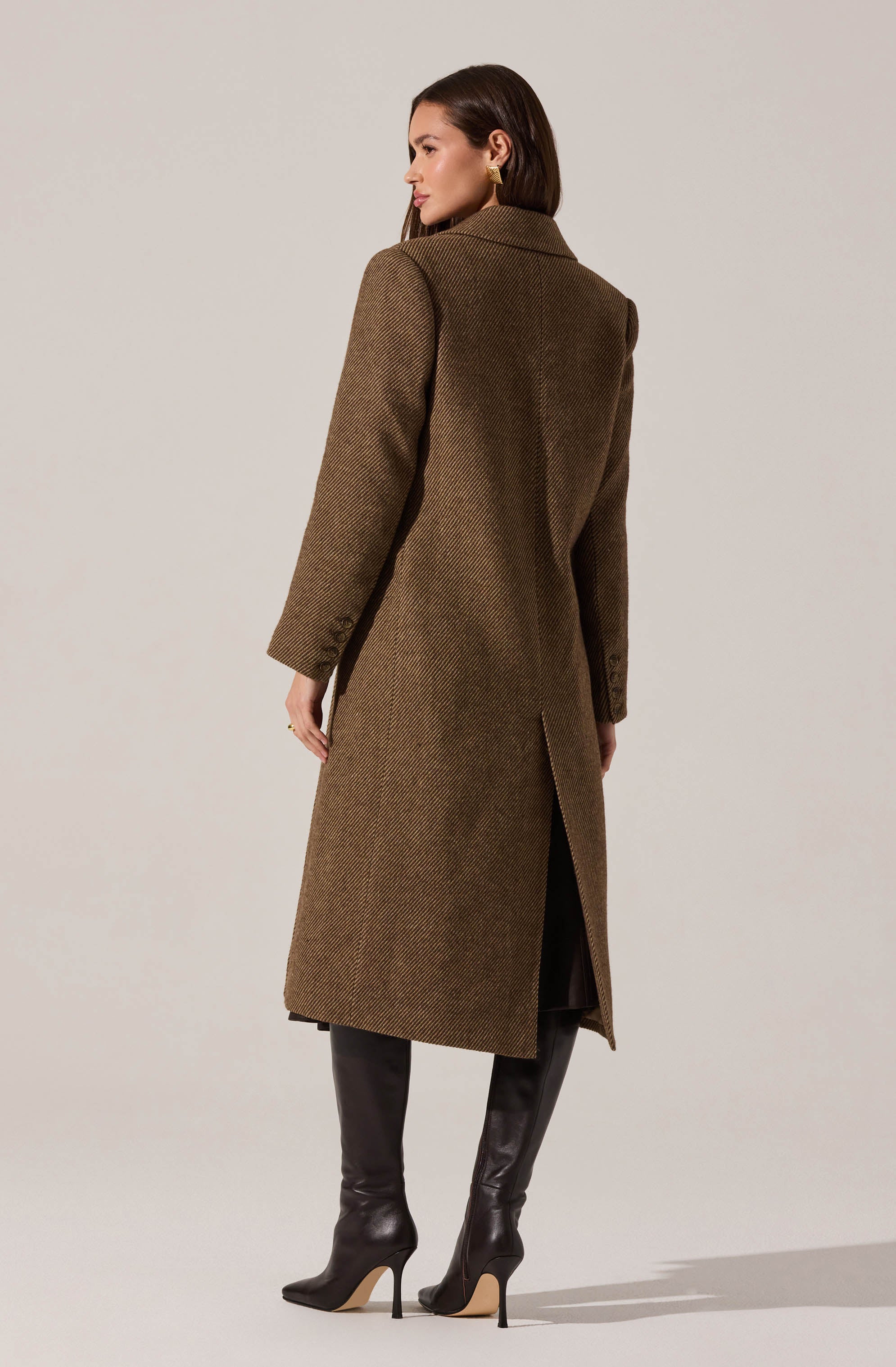 A woman is wearing a long, tailored brown coat with a subtle herringbone pattern, a classic collar, and a vent at the back. She accessorizes with gold hoop earrings and tall black leather boots with high heels.