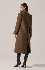 A woman is wearing a long, tailored brown coat with a subtle herringbone pattern, a classic collar, and a vent at the back. She accessorizes with gold hoop earrings and tall black leather boots with high heels.