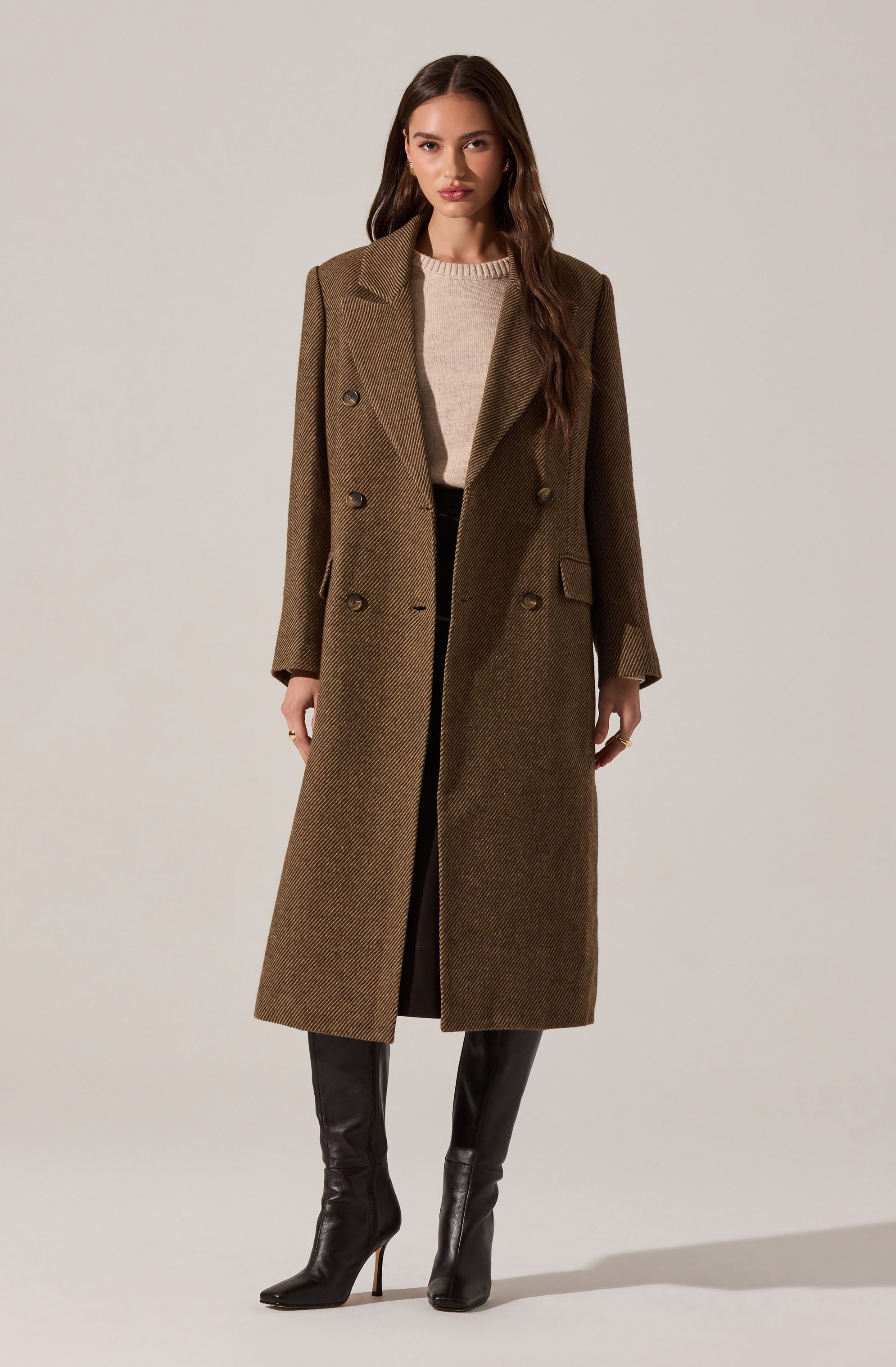 A woman is wearing a long, brown, double-breasted coat with a houndstooth pattern, featuring wide lapels and a structured fit. She has on a beige sweater underneath and black knee-high boots with high heels.