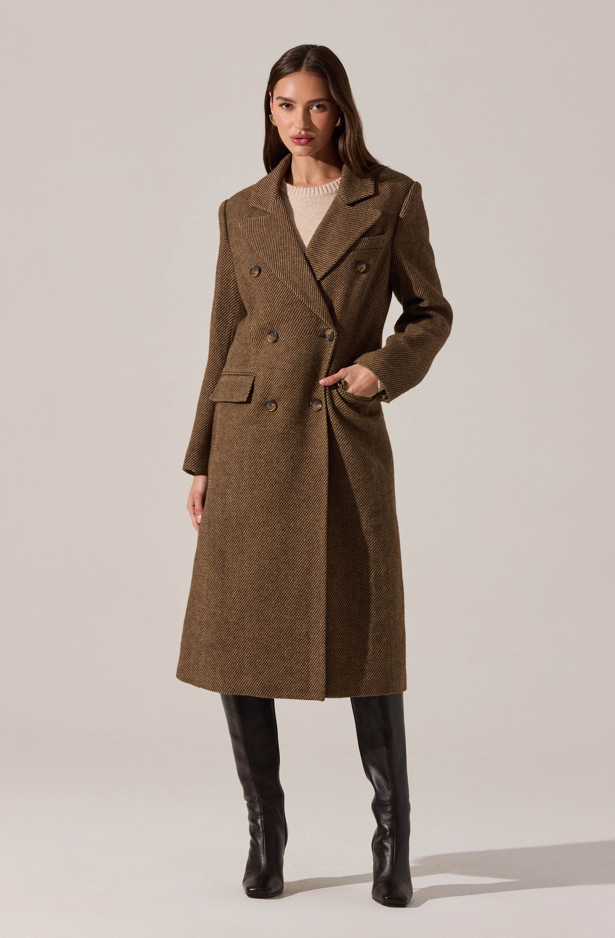 A woman is wearing a long, brown double-breasted coat with a textured, herringbone pattern. The coat features wide lapels, shoulder pads, and flap pockets. She is also dressed in black knee-high boots.
