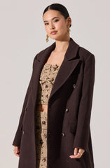 A woman is wearing a dark brown, double-breasted coat with wide lapels and buttons down the front. Underneath, she has on a matching beige, textured, cropped top and skirt set with a floral pattern. She accessorizes with large gold hoop earrings.