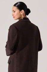 A woman with dark hair styled in a low bun is wearing a dark brown, textured blazer with a notched collar and long sleeves. She has large gold hoop earrings.