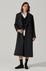 A woman is wearing a long, black pinstripe trench coat over a white collared shirt and a black cardigan. She has paired the outfit with black socks and black loafers. The coat features a classic lapel collar and double-breasted buttons.