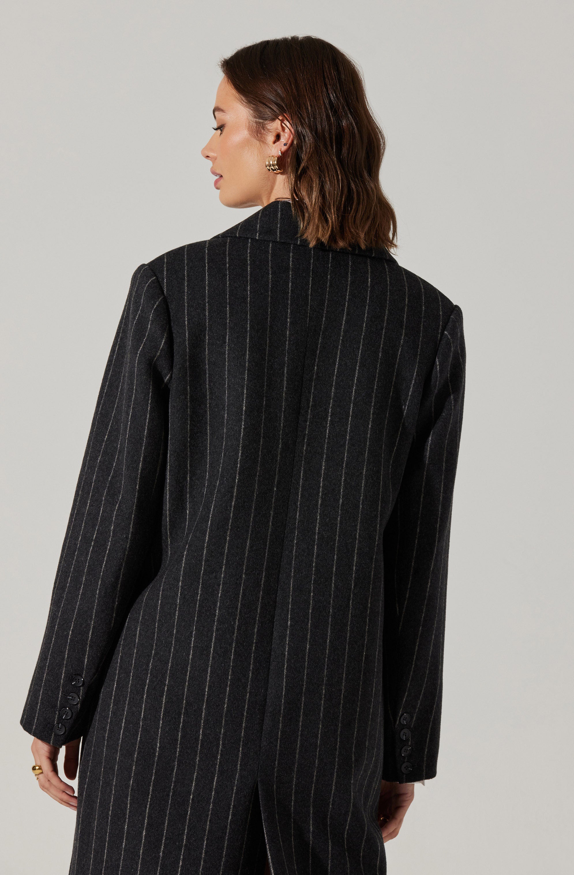 A woman is wearing a dark gray pinstripe blazer with light-colored vertical stripes. The blazer features a notched lapel collar and has buttons on the cuffs of the long sleeves. The back of the blazer extends down past her waist, with a slit at the center.