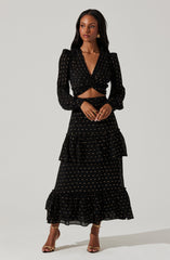 A woman is wearing a black two-piece outfit with a gold polka dot pattern. The top has long puffed sleeves, a V-neckline, and is twisted at the waist, exposing some midriff. The skirt is high-waisted, tiered with ruffles, and falls to her ankles.