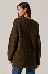 A woman is wearing a oversized, knit cardigan sweater in a dark brown color. The sweater features a textured cable knit pattern down the back and sleeves. She is also wearing blue jeans and has small hoop earrings.
