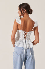 A woman is wearing a white eyelet lace top with ruffled shoulder straps and a back lace-up detail tied with a bow at the waist. The top has floral eyelet embroidery patterns and a ruffled hem. She is also wearing light-wash, high-waisted jeans with a relaxed fit.