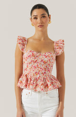 The woman is wearing a sleeveless, fitted top with ruffled shoulder straps. The top has a floral pattern in shades of pink, orange, and white, with a ruched bodice and a peplum-style ruffle at the hem. She is also wearing a pair of white pants.